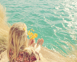 Menina olha o mar com flor na mão, sentada em uma pedra